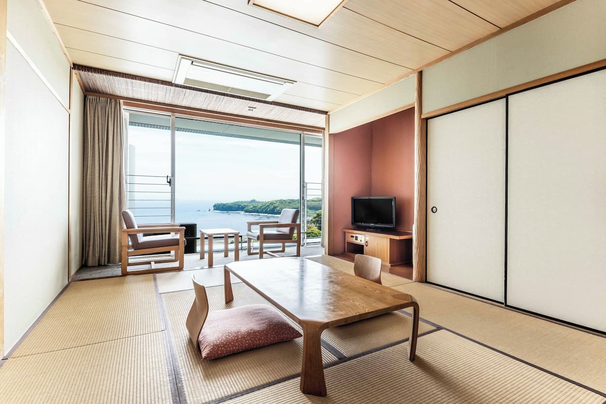 Une pièce avec un sol recouvert de tatamis, une table en bois, des chaises, un téléviseur et une porte coulissante.