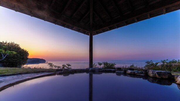 Une piscine à débordement paisible, offrant une vue dégagée sur le coucher de soleil sur l’eau, et entourée de rochers et de verdure.