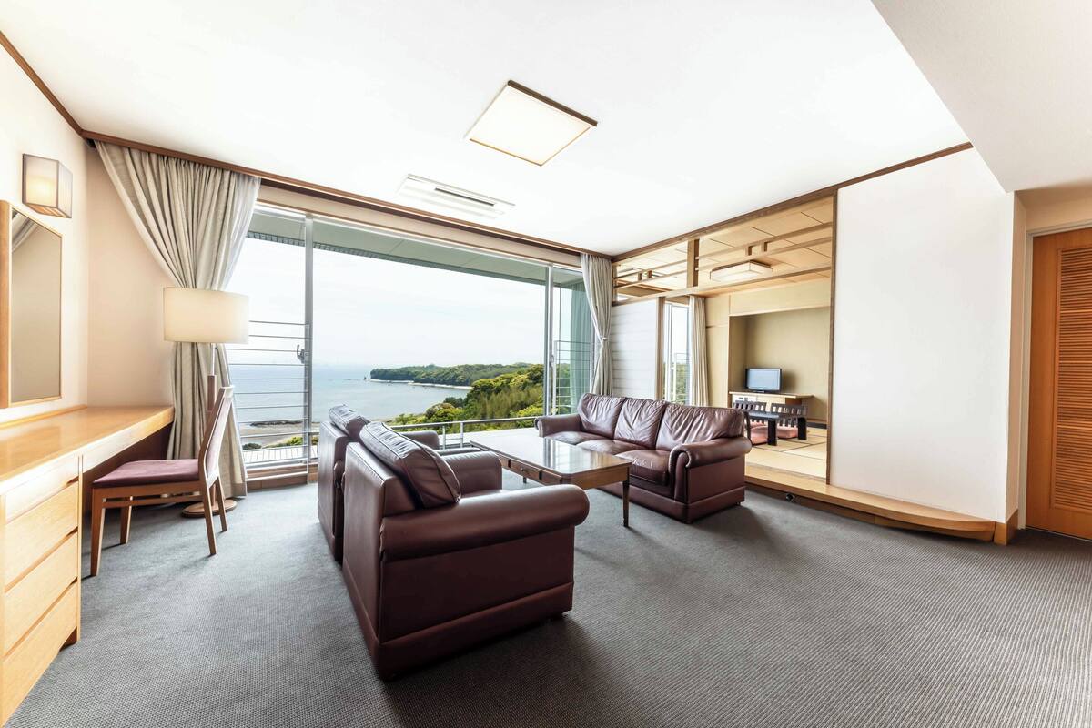 Une chambre d’hôtel moderne dotée d’une grande fenêtre offrant une vue panoramique, d’un coin salon avec des canapés en cuir, d’un bureau en bois et d’un téléviseur à écran plat.