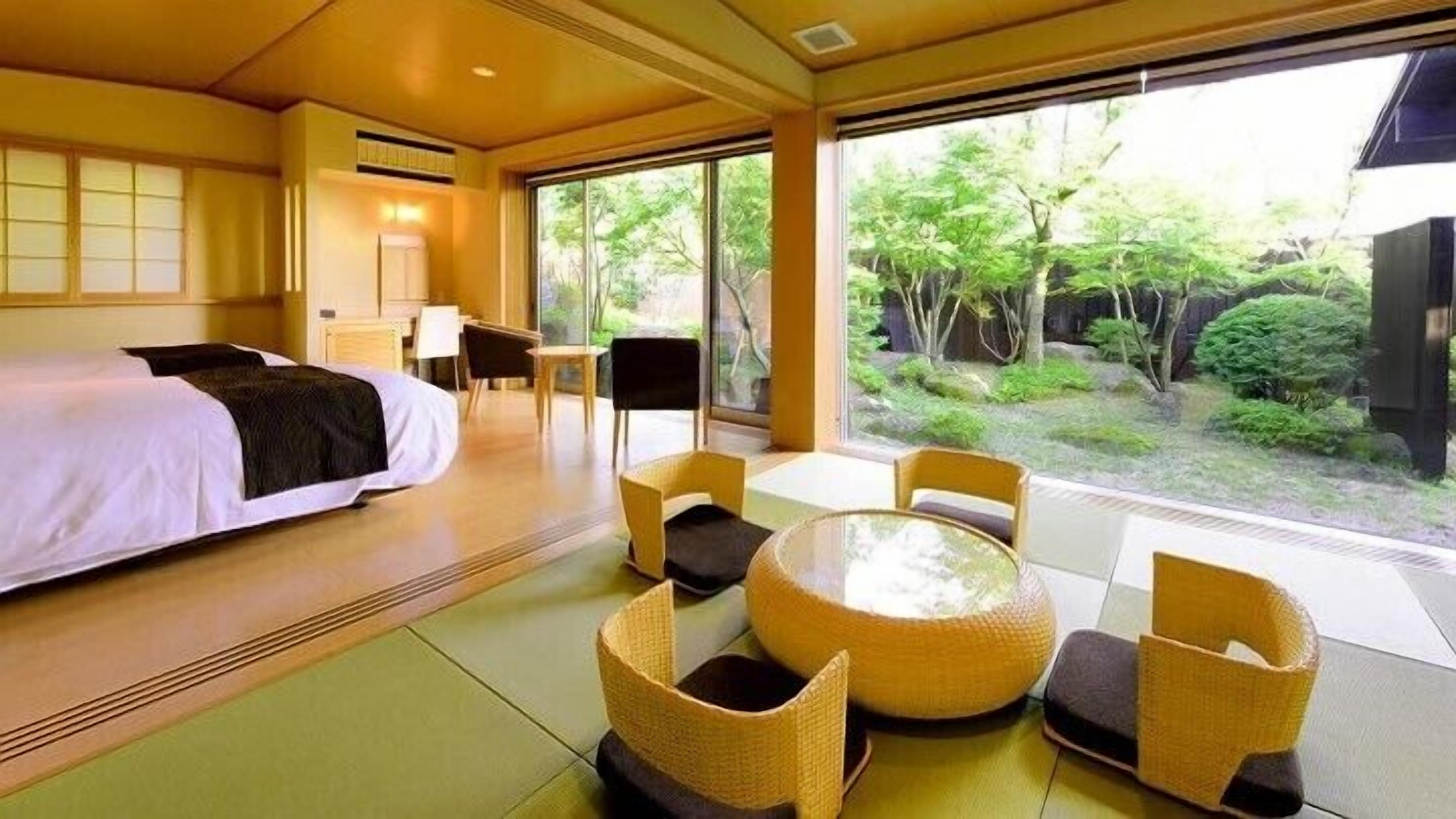 A hotel room with a bed, a dining table with chairs, and a view of greenery outside.