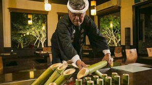 A person arranging bamboo shoots on a table in a room with large windows and indoor plants. - Okuyado Buaisou (Yufu)