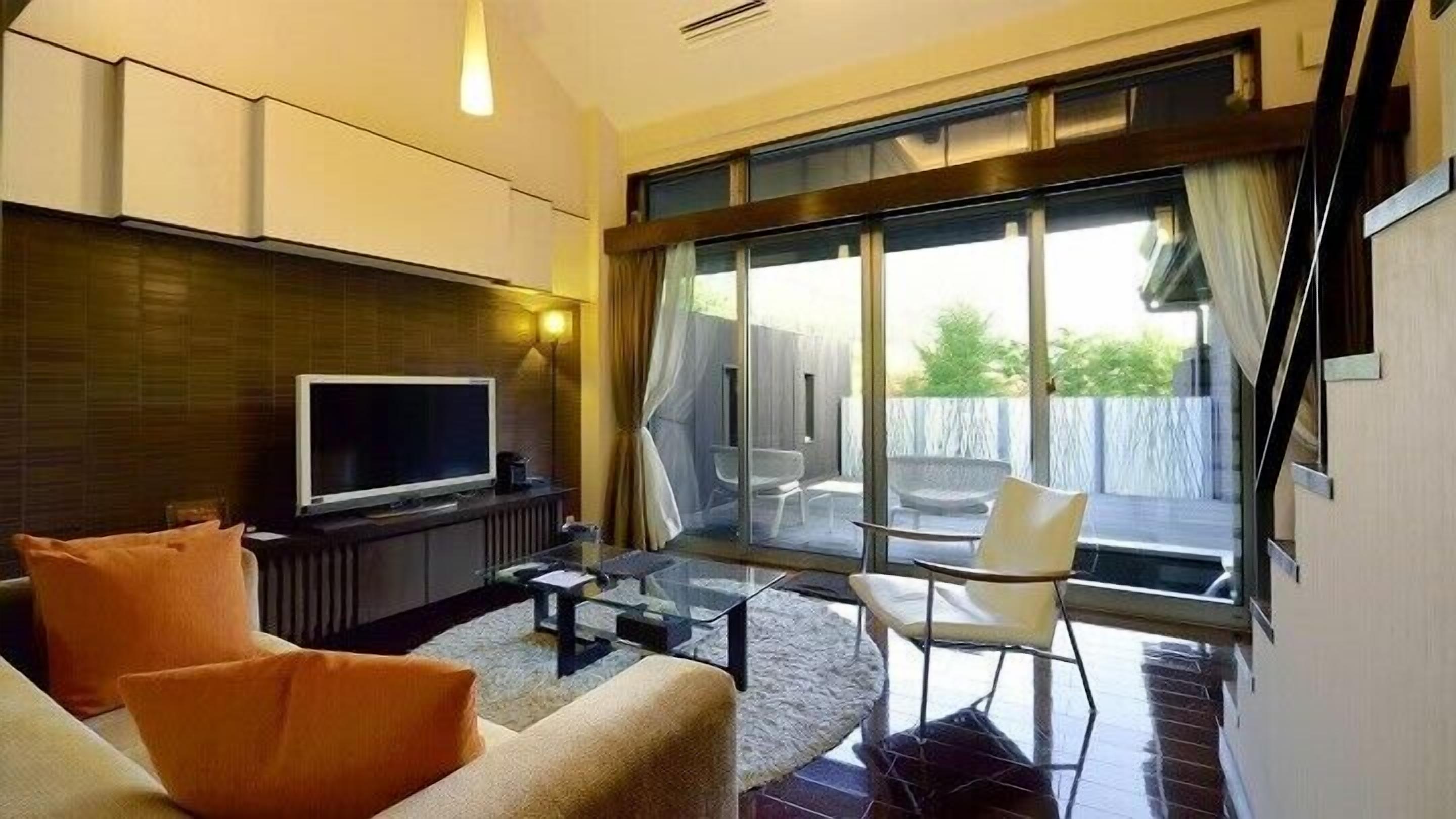 A modern living room with a flat-screen TV, sofa, and chairs, a glass door leading to an outdoor area with a view.