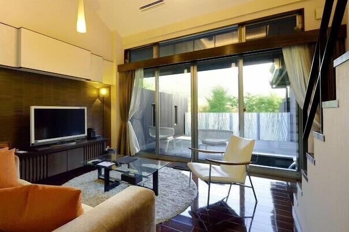 A modern living room with a flat-screen TV, sofa, and chairs, a glass door leading to an outdoor area with a view.