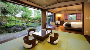 Habitació amb paviment de tatami, una taula baixa, dues cadires amb coixins, un llit i vista a un jardí amb roques i vegetació.