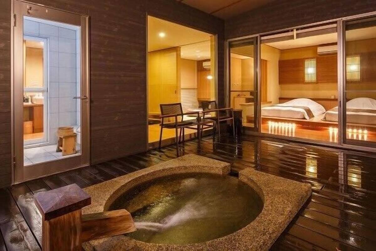 A room with a hot tub, a wooden floor, a bed with white bedding, and a view through a glass door.
