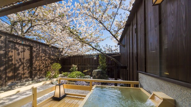 A traditional Japanese onsen with a wooden deck, a stone wall, and cherry blossoms in the background. - Inisienoyado Keiunn (Izumo)