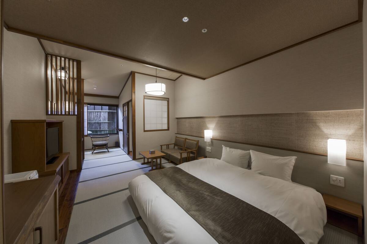 A hotel room with a large bed, bedside tables, a TV, a seating area with a table and chairs, and a view of a traditional Japanese room.