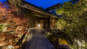 A traditional Japanese building with a stone pathway leading to a wooden bridge over a small pond, surrounded by foliage and stone lanterns. - Inisienoyado Keiunn (Izumo)