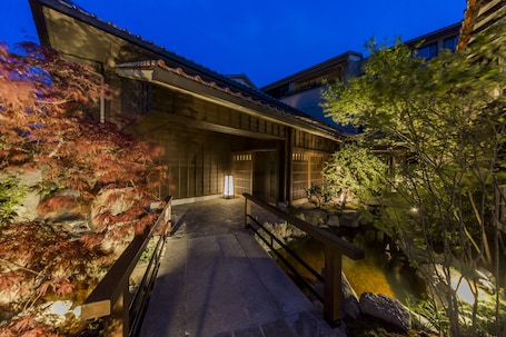 A traditional Japanese building with a stone pathway leading to a wooden bridge over a small pond, surrounded by foliage and stone lanterns.