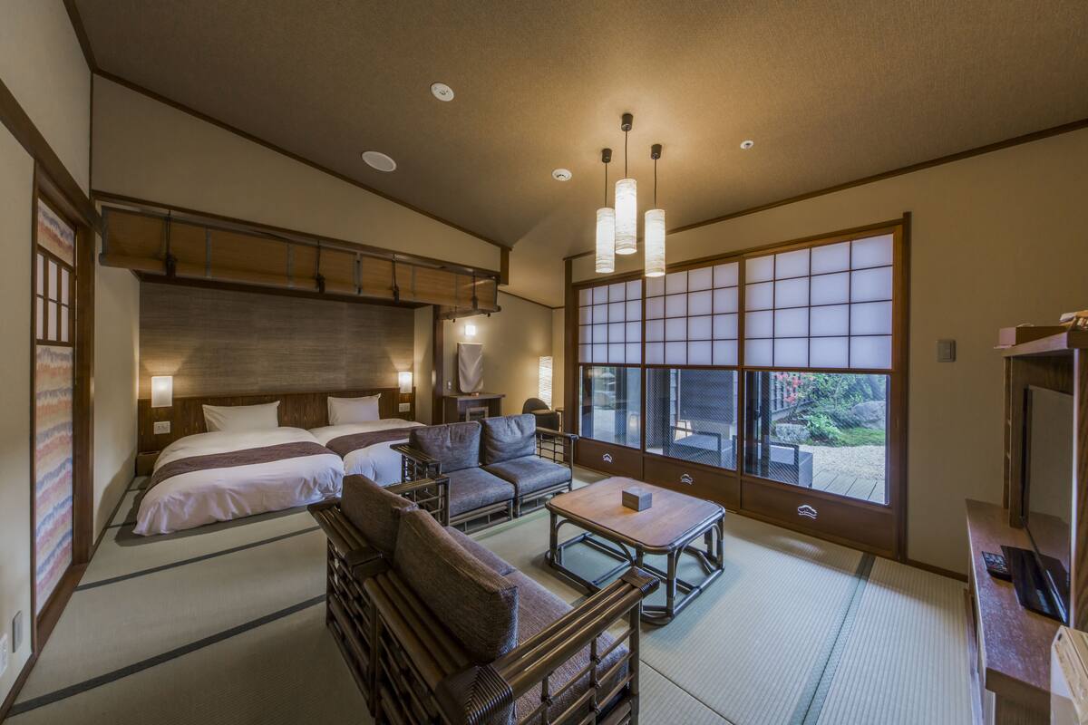 A traditional Japanese-style hotel room with tatami flooring, a futon bed, a sofa, a small table, and a sliding shoji screen window.