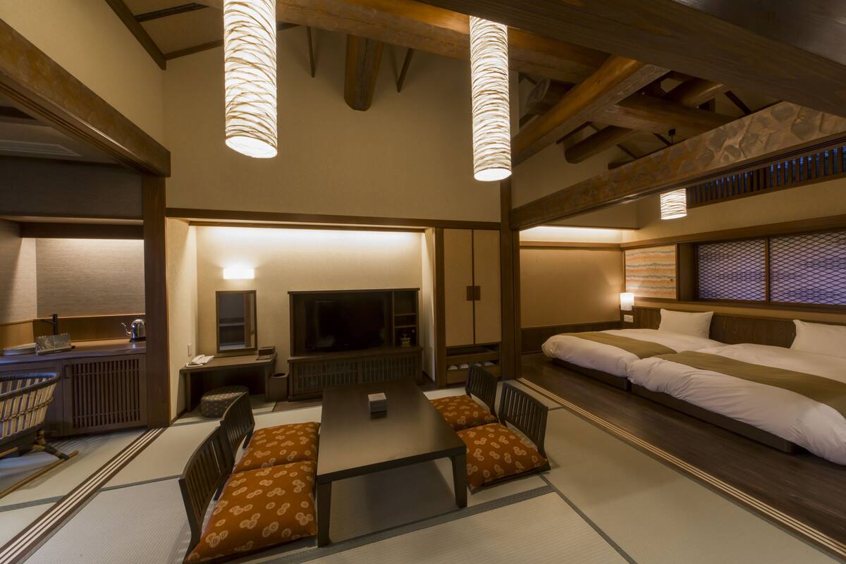 A traditional Japanese-style room with tatami flooring, a low table, cushions, a television, and a bed with a futon.