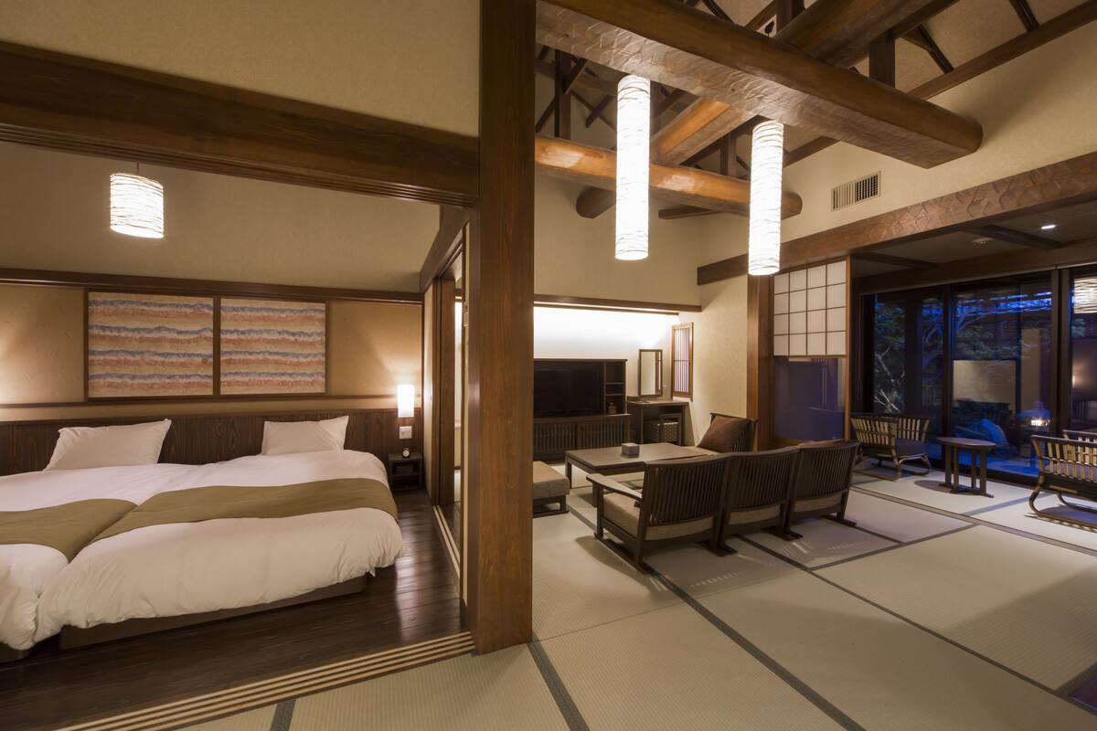 A traditional Japanese-style hotel room with tatami flooring, a large bed, a sitting area with low tables and benches, and a view of the outside.