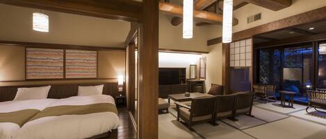 A traditional Japanese-style hotel room with tatami flooring, a large bed, a sitting area with low tables and benches, and a view of the outside.