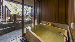 A traditional Japanese onsen with a large wooden bathtub, stone steps leading to a natural water feature, and a view of a wooden building outside. - Inisienoyado Keiunn (Izumo)