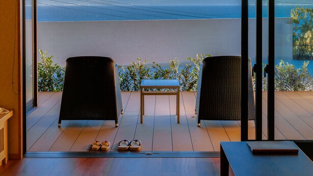 A room with a view of a body of water and mountains, two wicker chairs, a small table, and a pair of shoes on the floor. - Futaritowani En (Izu)
