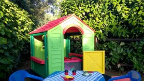 Une maison de jeu pour enfants, avec un toit rouge et des murs verts, entourée d’une végétation luxuriante.