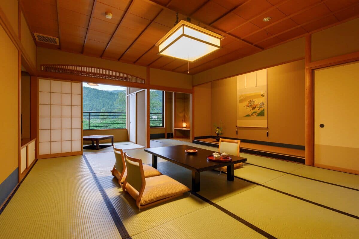 A traditional Japanese room with tatami flooring, a low table, cushions, sliding doors, and a view of greenery.