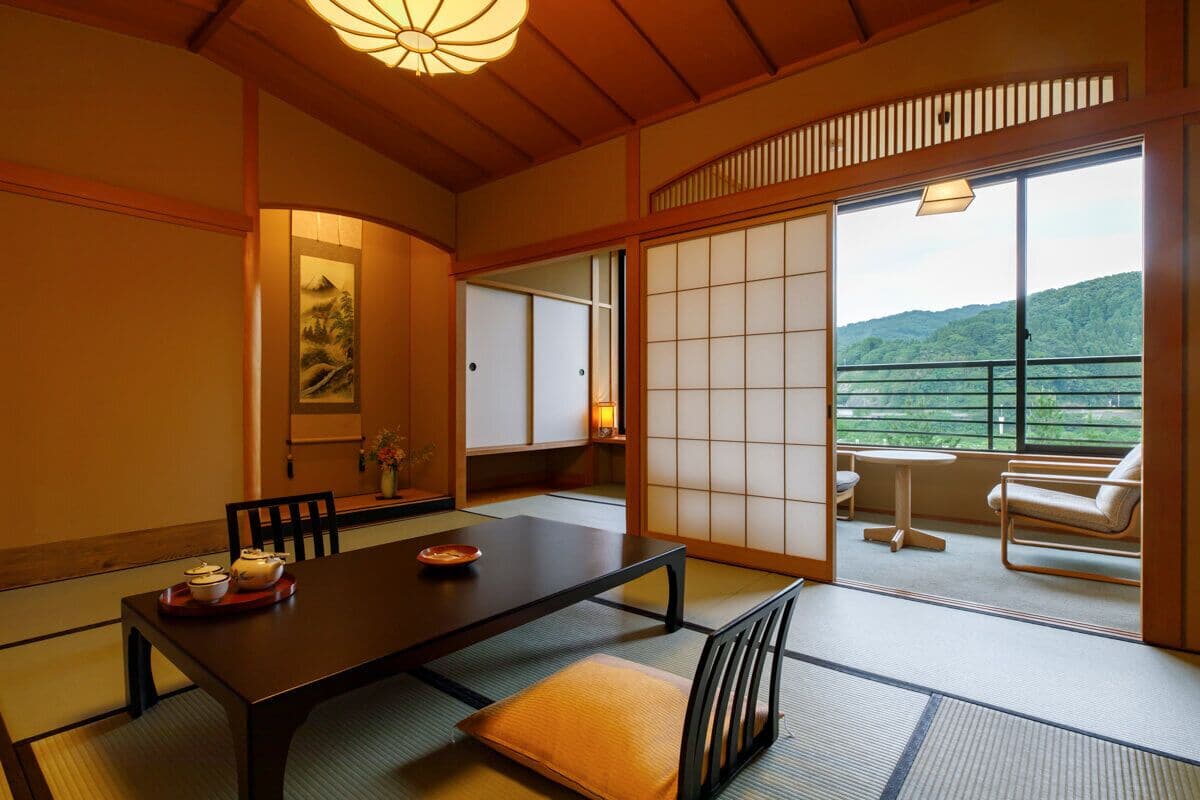 A traditional Japanese room with tatami flooring, a low table, chairs, sliding doors, and a balcony with a view of the hills.