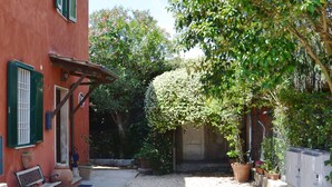 Un patio con un edificio rojo, contraventanas verdes, un banco y macetas con plantas.