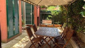 Un porche cubierto con una mesa y sillas, rodeado de vegetación y un muro de ladrillo.