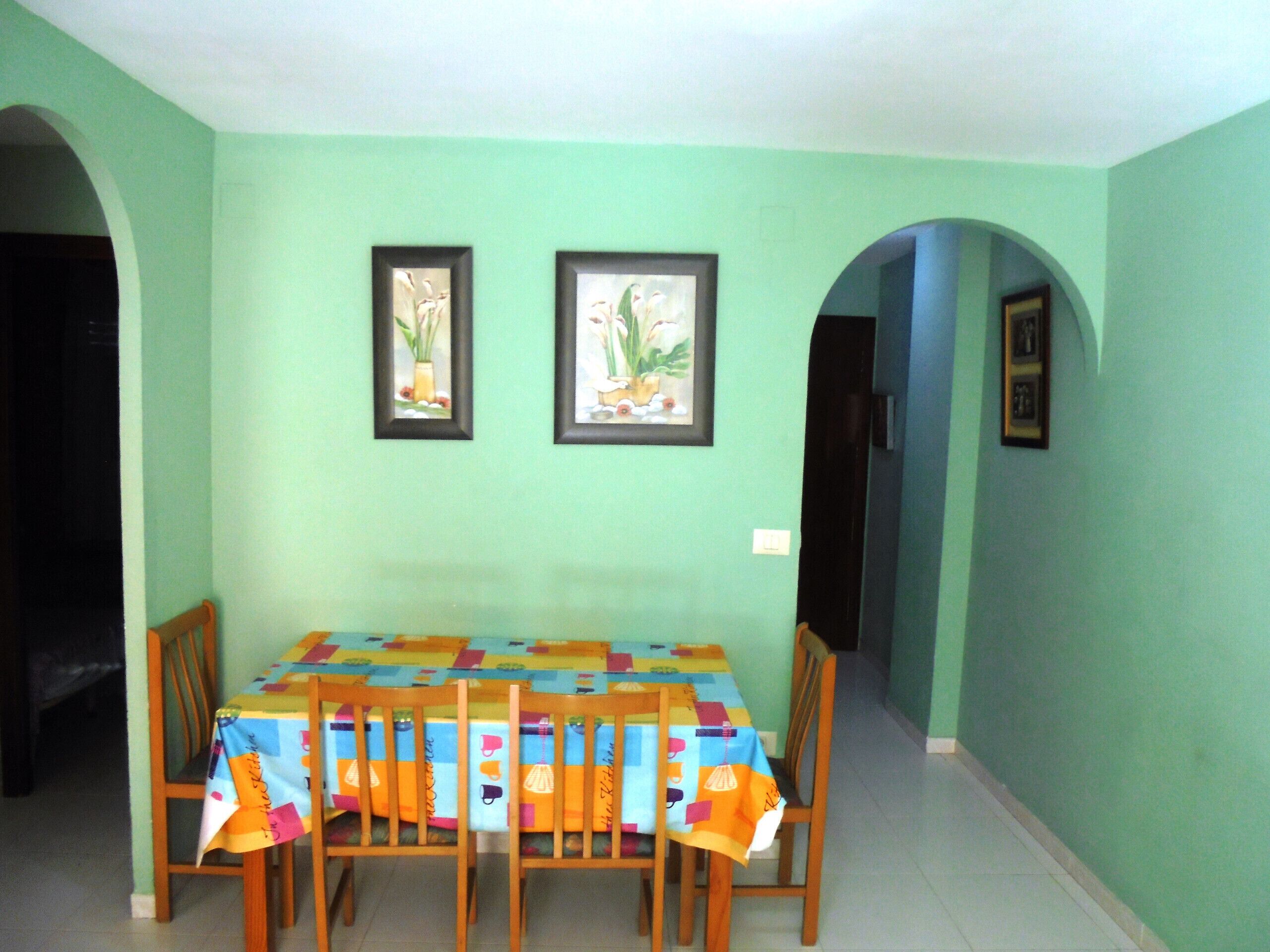 Un coin repas avec une table recouverte d’une nappe colorée, des chaises en bois et deux tableaux encadrés accrochés au mur.