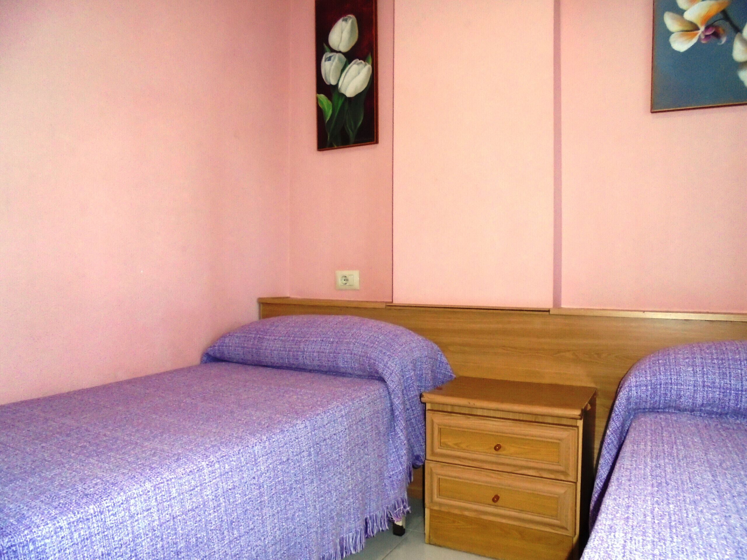 A bedroom with two single beds, a wooden nightstand, and a wall-mounted light switch.