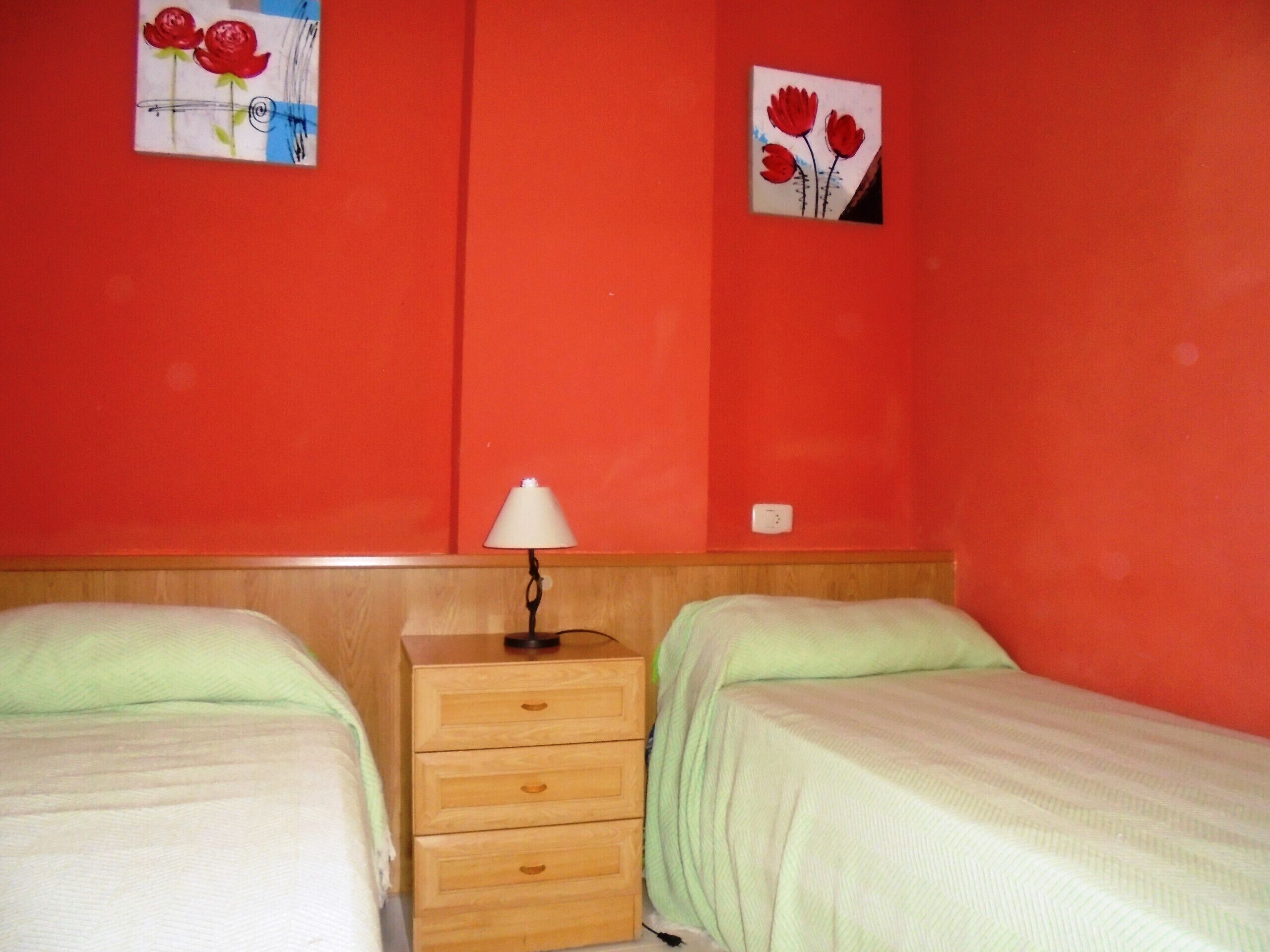 A room with two beds, a wooden nightstand with a lamp, and two wall-mounted paintings of flowers.