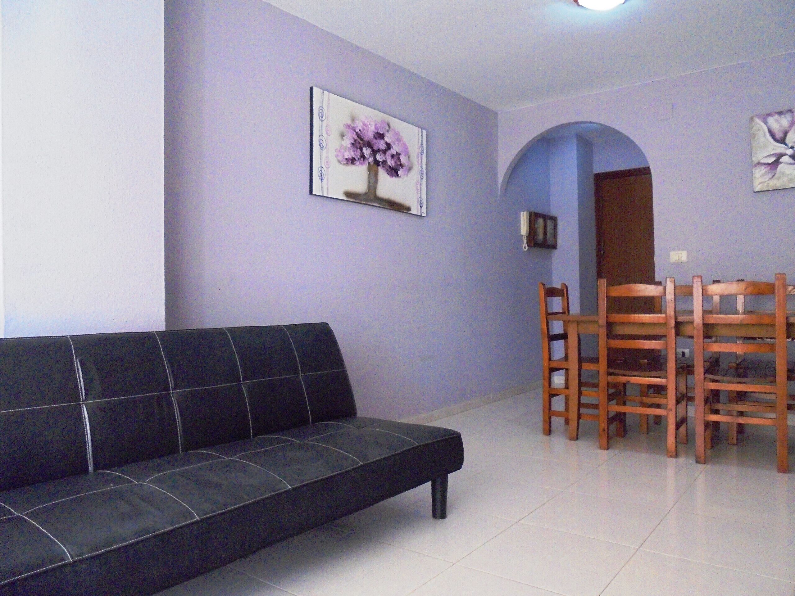 Ein Wohnzimmer mit einem schwarzen Sofa, einem Esstisch mit Holzsesseln und einer violetten Wand mit einem gerahmten Bild.