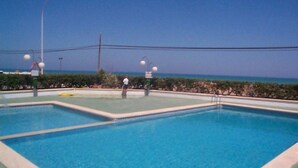 Una piscina rectangular con agua azul cristalina, rodeada por un muro bajo de color verde y un borde blanco. Se observan dos escaleras metálicas en el borde de la piscina, y algunas personas al fondo.