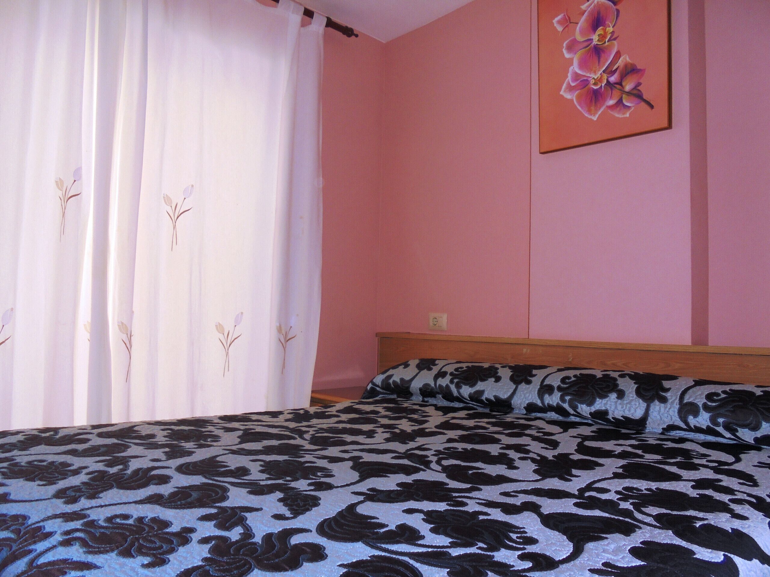 A bedroom with a bed, a window with sheer curtains, and a wall with a floral painting.