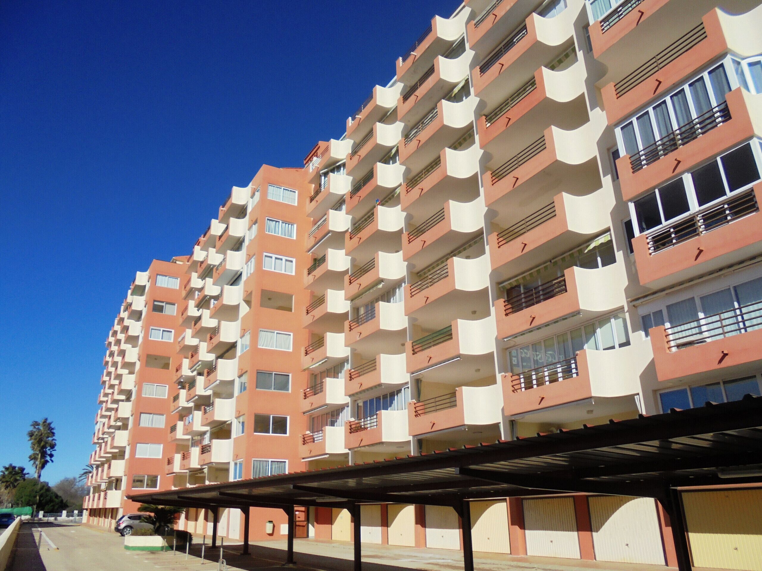 A multi-story residential building with balconies, a covered parking area, and a clear blue sky.