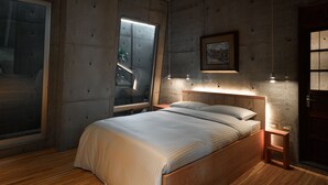 A bedroom with a large bed, a nightstand, a wall-mounted light, and a window with blinds. - Mao House (Tainan)