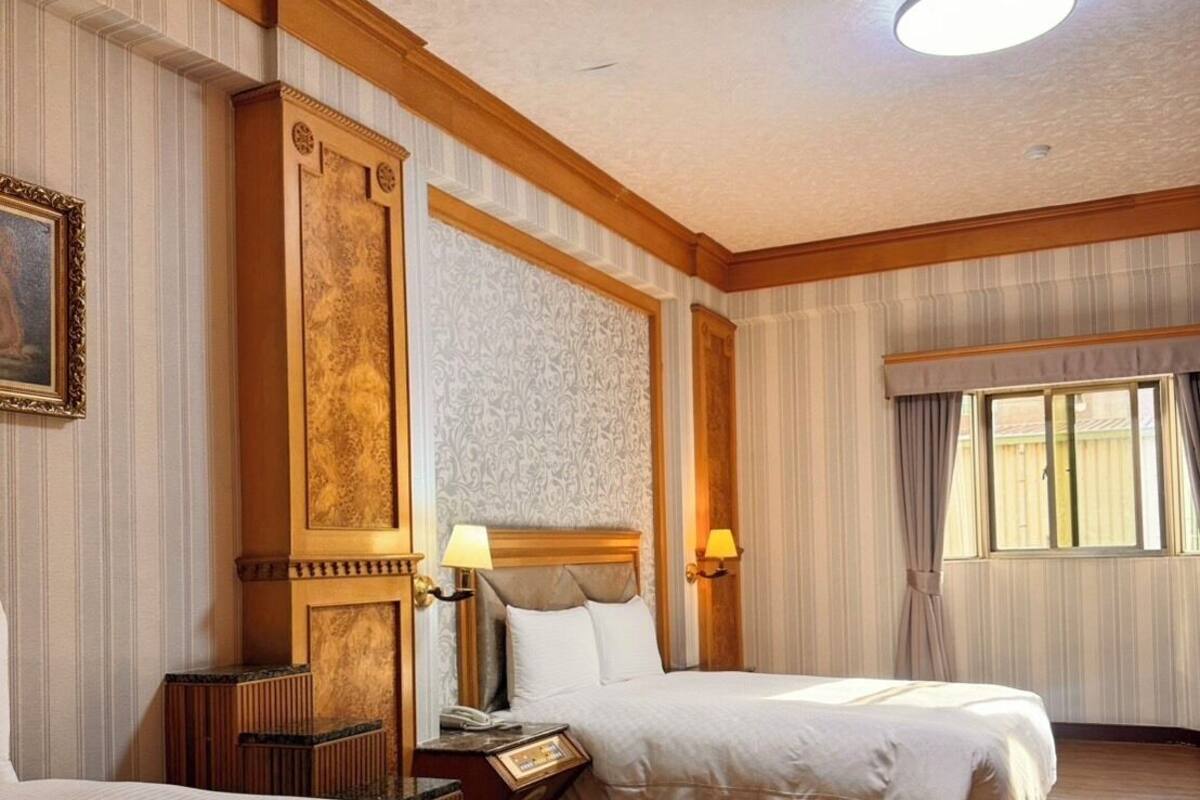 A hotel room with two beds, a wooden headboard, a nightstand, a lamp, and a window with curtains.