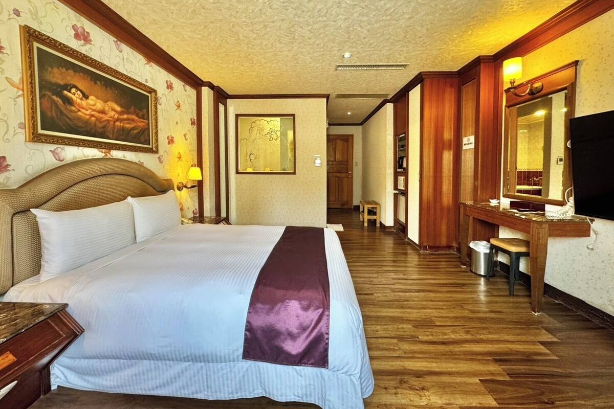 A hotel room with a large bed, wooden flooring, a television, a desk with a mirror, and a bathroom visible through an open door.