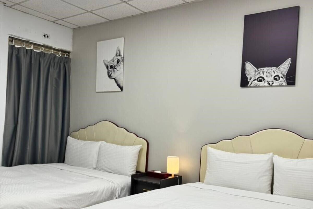 A hotel room with two beds, a nightstand with a lamp, and two wall art pieces of cats.