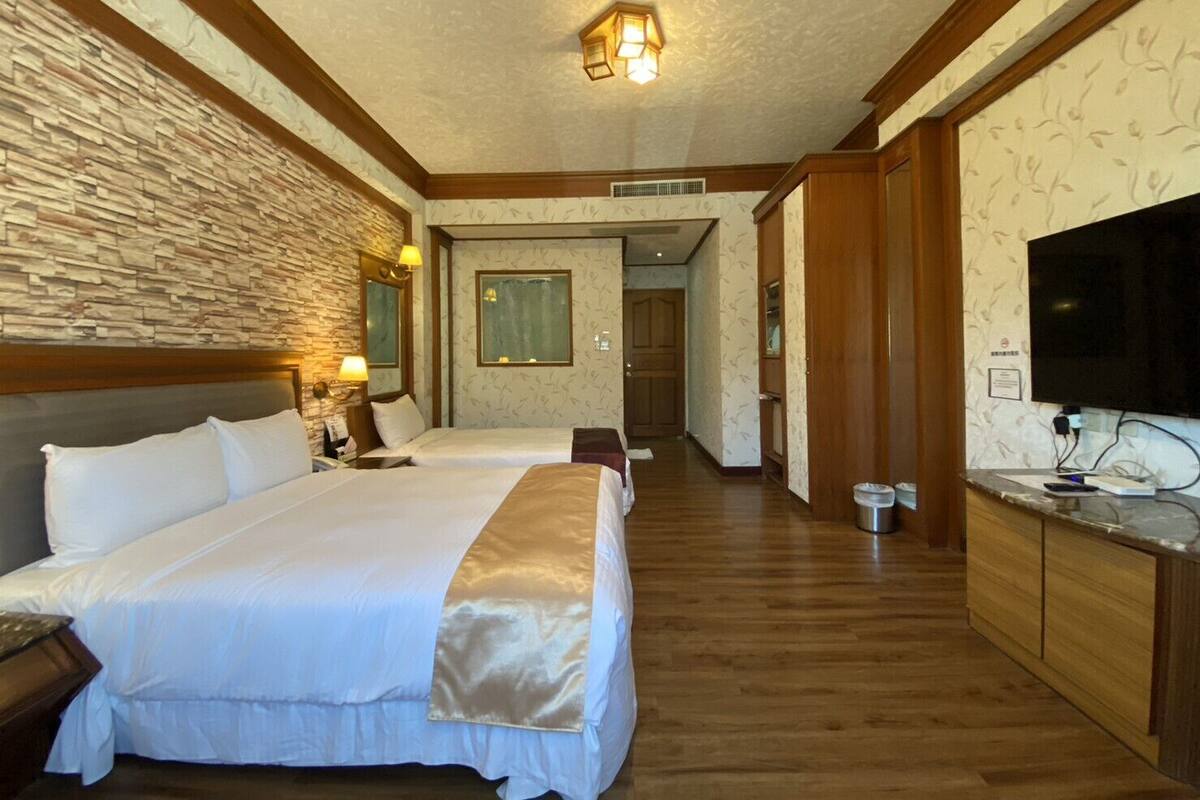 A hotel room with two beds, a stone accent wall, wooden flooring, a TV, and a small kitchenette.