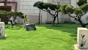 Un jardin paisible, avec des arbustes soigneusement taillés, un banc en pierre et un monument en pierre orné de motifs floraux.