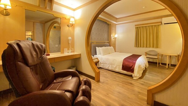 Une chambre d’hôtel équipée d’un fauteuil massant, d’un lit avec une couverture rouge, d’une tête de lit en bois, d’une petite table et d’un miroir.