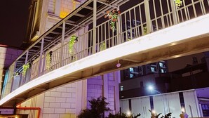 Un pont piétonnier doté de garde-corps et orné de plantes suspendues, qui mène à un bâtiment surmonté d’une tour horloge.
