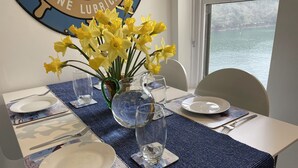 Una mesa de comedor dispuesta con platos, cubiertos y un jarrón de flores amarillas, con vista a una masa de agua a través de una ventana.