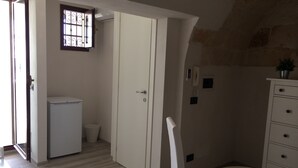 Een kleine kamer met een bed, een tafeltje met een afstandsbediening, een witte kledingkast, een deur naar een keuken en een raam met tralies.
