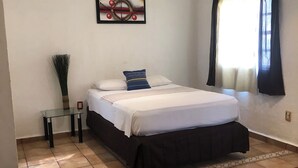 Un dormitorio con una cama, una mesita, una silla, una ventana con cortinas y un cuadro colgado en la pared.