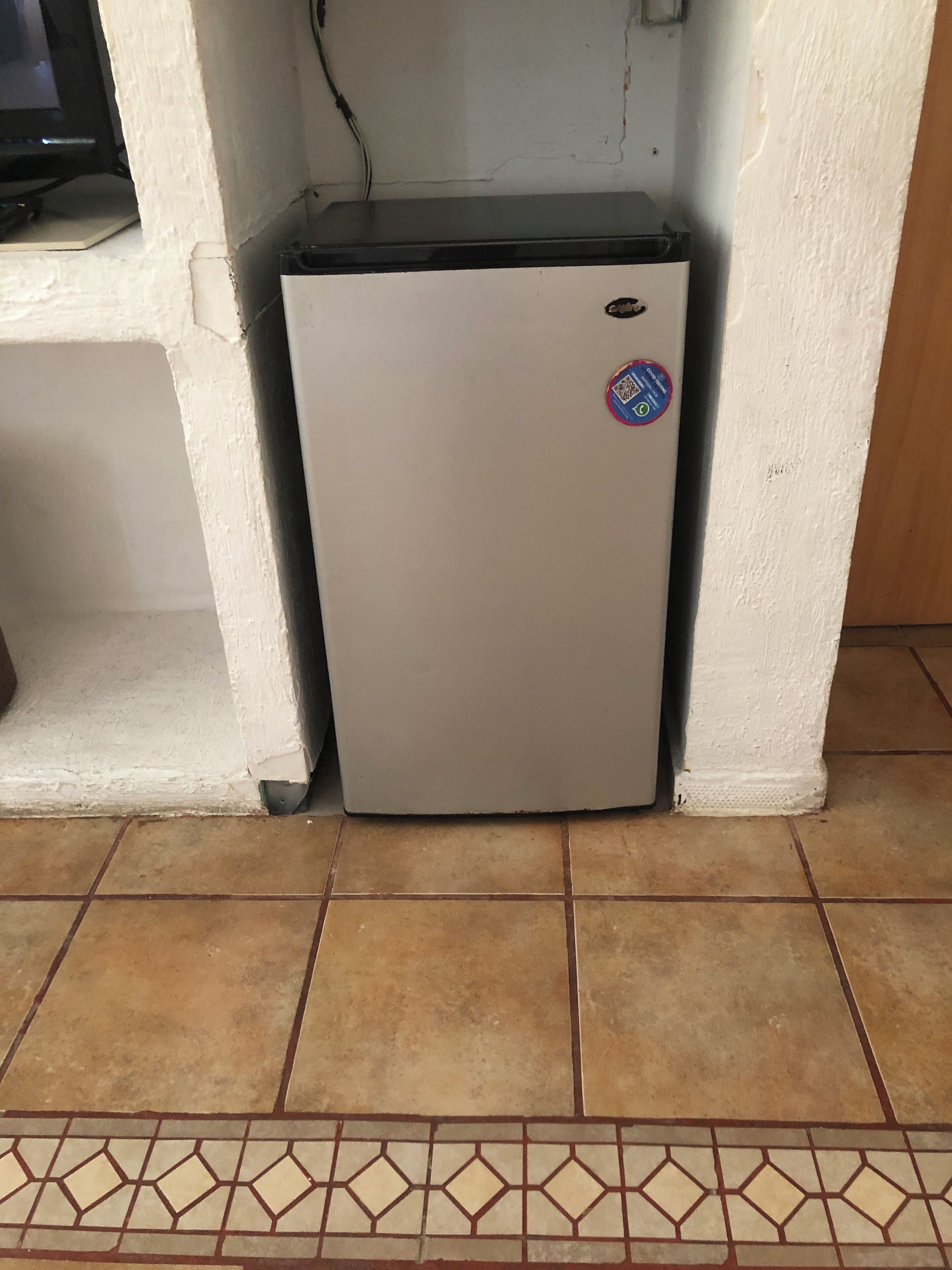 A small, white refrigerator with a QR code sticker, located in a corner with tiled flooring and a white wall.