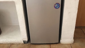 Un refrigerador pequeño y blanco con un adhesivo con código QR, situado en una esquina con piso de baldosas y una pared blanca.