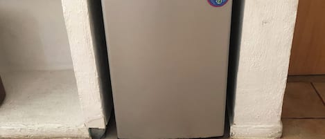 A small, white refrigerator with a QR code sticker, located in a corner with tiled flooring and a white wall.