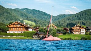 Een dorp aan het meer met traditionele houten huizen, een zeilboot en beboste heuvels op de achtergrond.