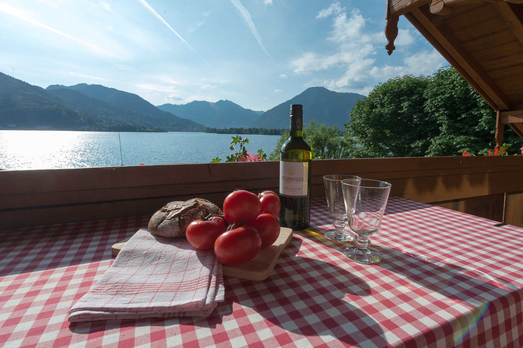 Una mesa con mantel a cuadros, una botella de vino, copas, pan y tomates sobre una terraza de madera con vistas a un lago y montañas al fondo.