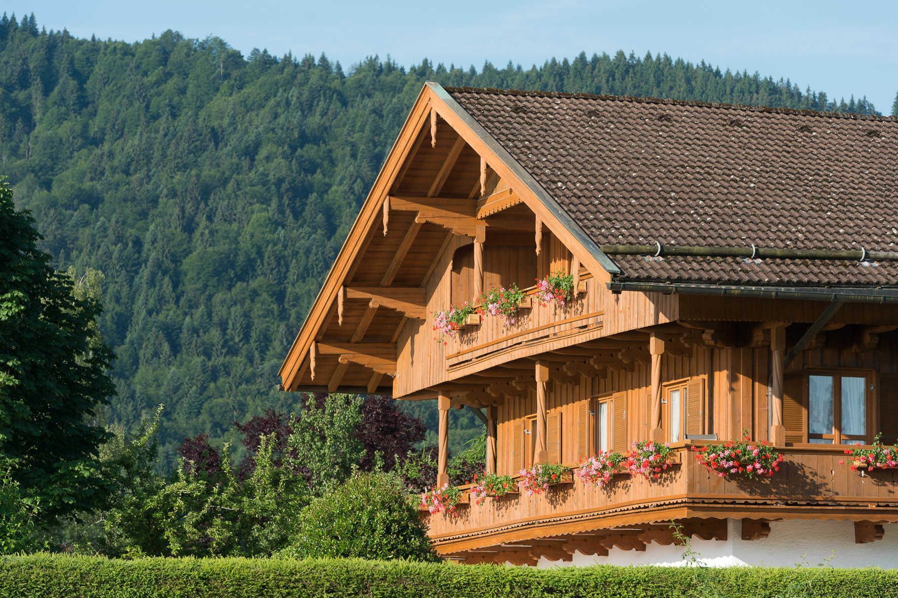 A wooden chalet with a balcony, surrounded by lush greenery and a forested mountain backdrop.