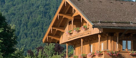A wooden chalet with a balcony, surrounded by lush greenery and a forested mountain backdrop.