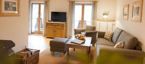 A living room with a sofa, ottoman, wooden table, television, and a window with curtains. - Vacation apartment 5 - Seerose am See - Dream vacation directly on Lake Tegernsee (Bad Wiessee)
